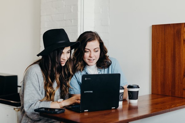 Two women sitting at a table with coffee cups while looking at a laptop.